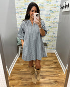 A woman takes a mirror selfie wearing a blue and white vertically striped short-sleeve tunic dress with tan suede boots in a fitting room with wood floors and abstract wallpaper.