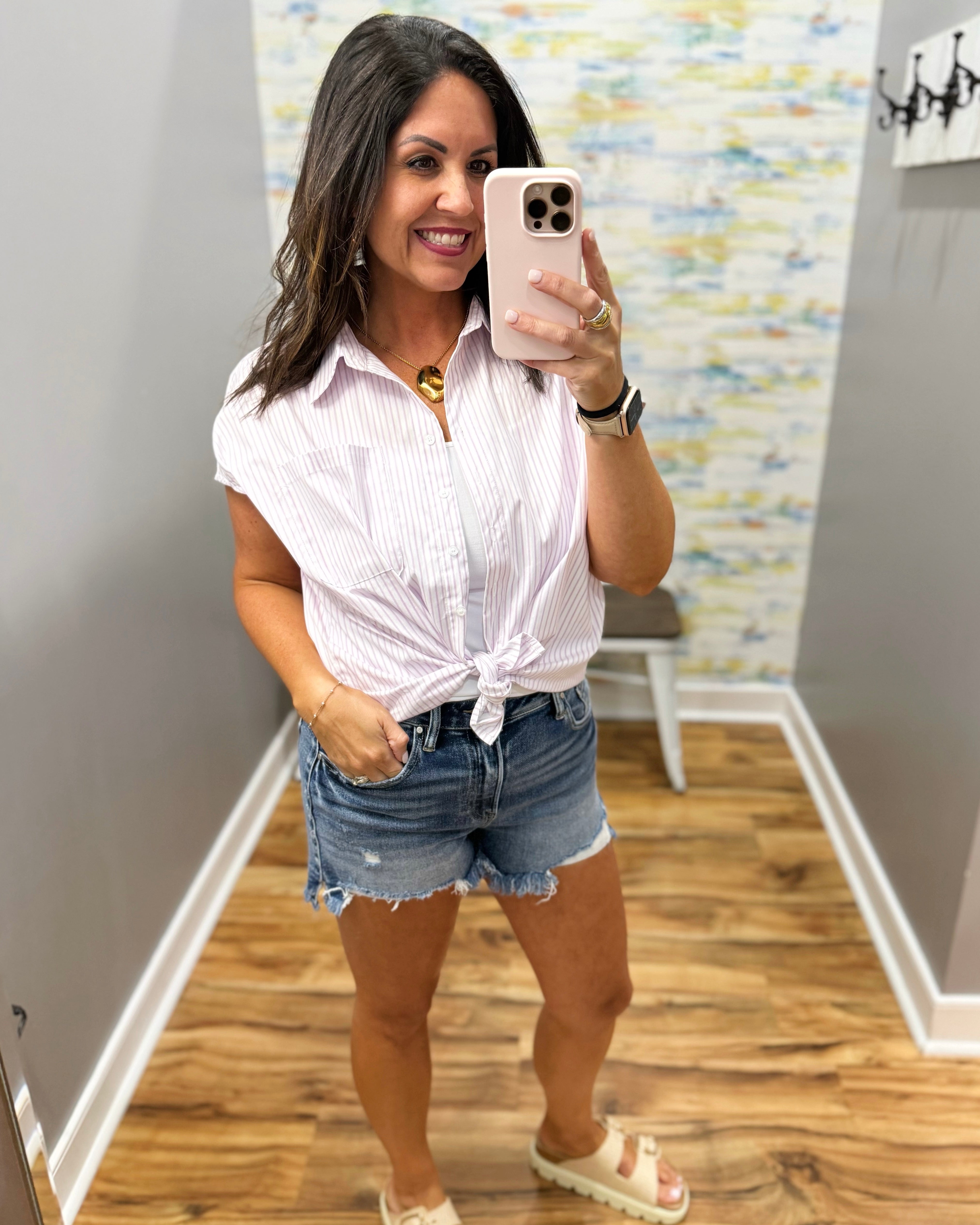 Woman taking a mirror selfie wearing a white striped shirt and denim shorts in a room with patterned wallpaper.