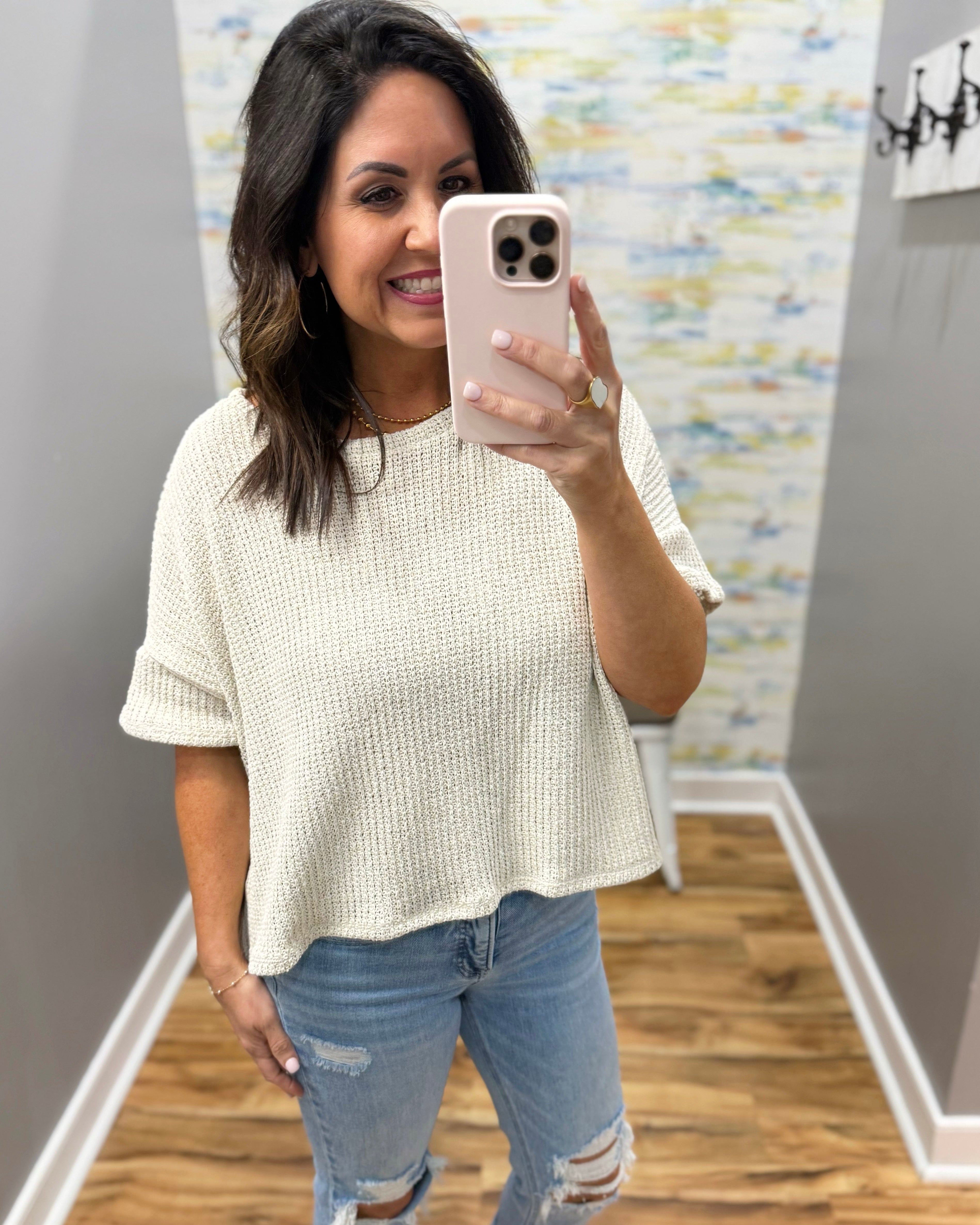Woman taking a mirror selfie wearing a natural sweater and jeans in a room with a textured wall.