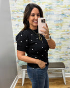 A woman takes a mirror selfie wearing a black short-sleeve knit top with white polka dots and blue jeans, standing in front of a patterned wall.