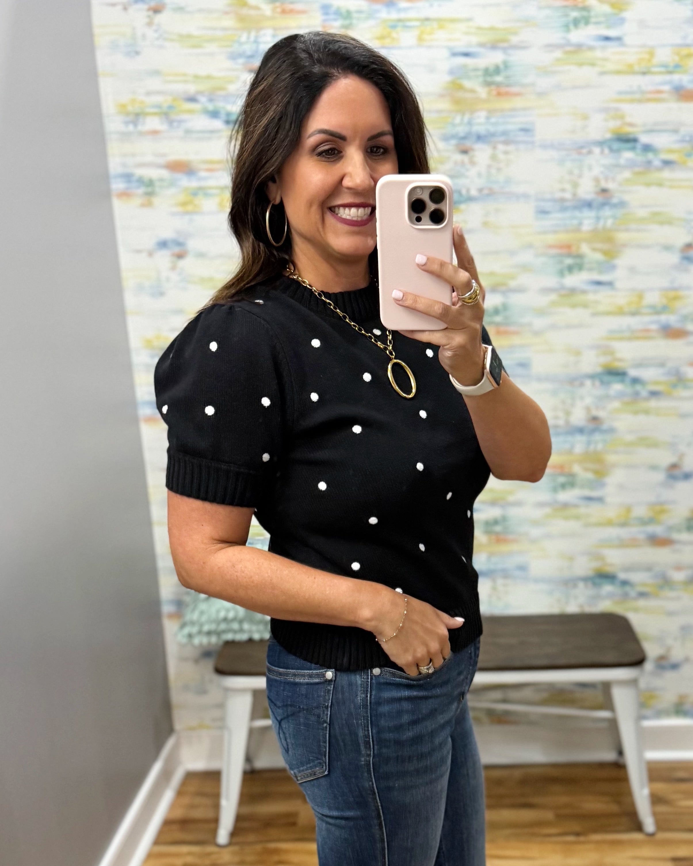 A woman takes a mirror selfie wearing a black short-sleeve knit top with white polka dots and blue jeans, standing in front of a patterned wall.
