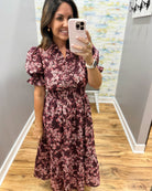 A woman takes a mirror selfie in a boutique wearing a dark burgundy maxi dress with a pink and white floral pattern, puff sleeves, and a smocked waist.