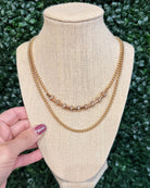 A gold-toned double-strand necklace displayed on a beige fabric jewelry bust, with a hand holding the left side of the bust. The necklace features two chains with a central row of small silver-toned square links.