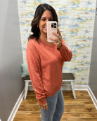 A woman takes a mirror selfie wearing a long-sleeve V-neck knit sweater in a muted terracotta or burnt orange color, paired with light blue jeans. The setting is a home fitting room with a wood floor, gray walls, and a colorful abstract wallpaper.