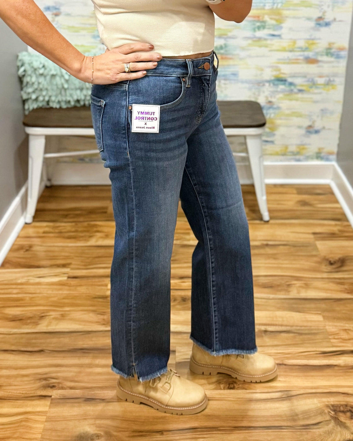 A person wearing blue Risen crop straight-leg jeans with a raw frayed hem, paired with beige boots and a cream top, standing on a wood floor in front of a white bench.