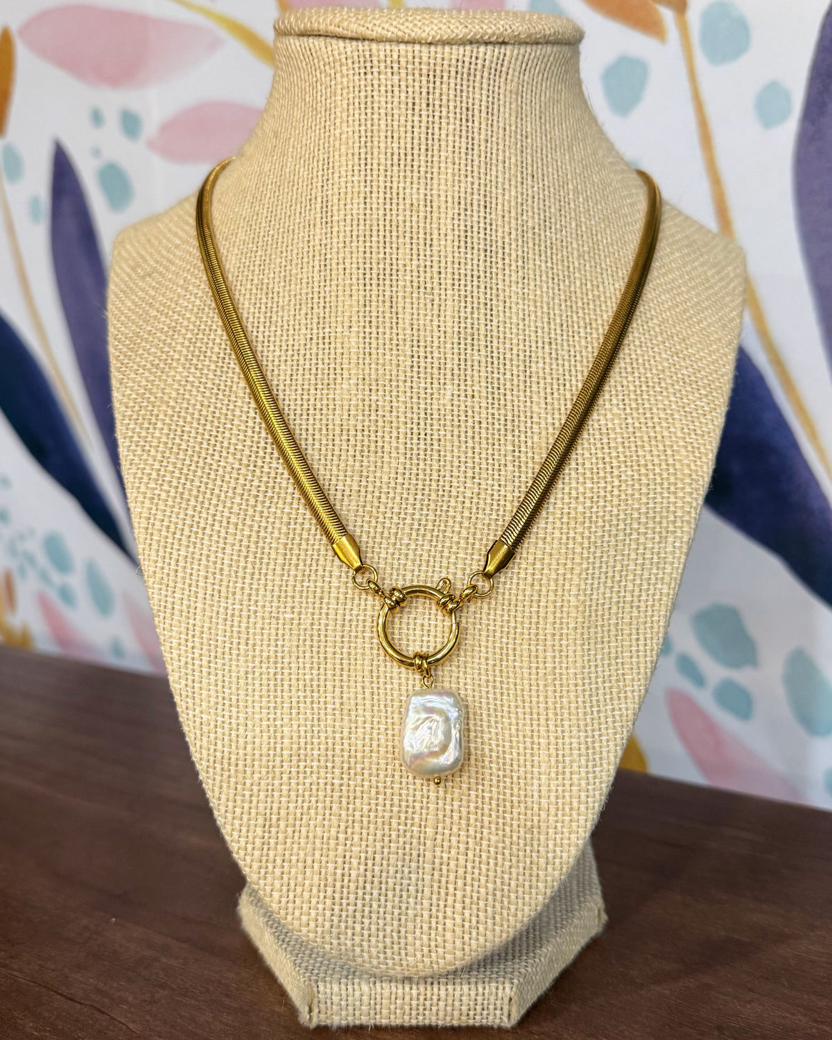 Chansuttpearls Delilah gold chain necklace with a toggle clasp and a small white carved pendant, displayed on a beige woven jewelry bust against a colorful patterned backdrop.