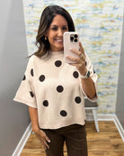 A woman taking a mirror selfie wearing a cream-colored knit sweater with large black polka dots and brown pants, holding a light pink smartphone.