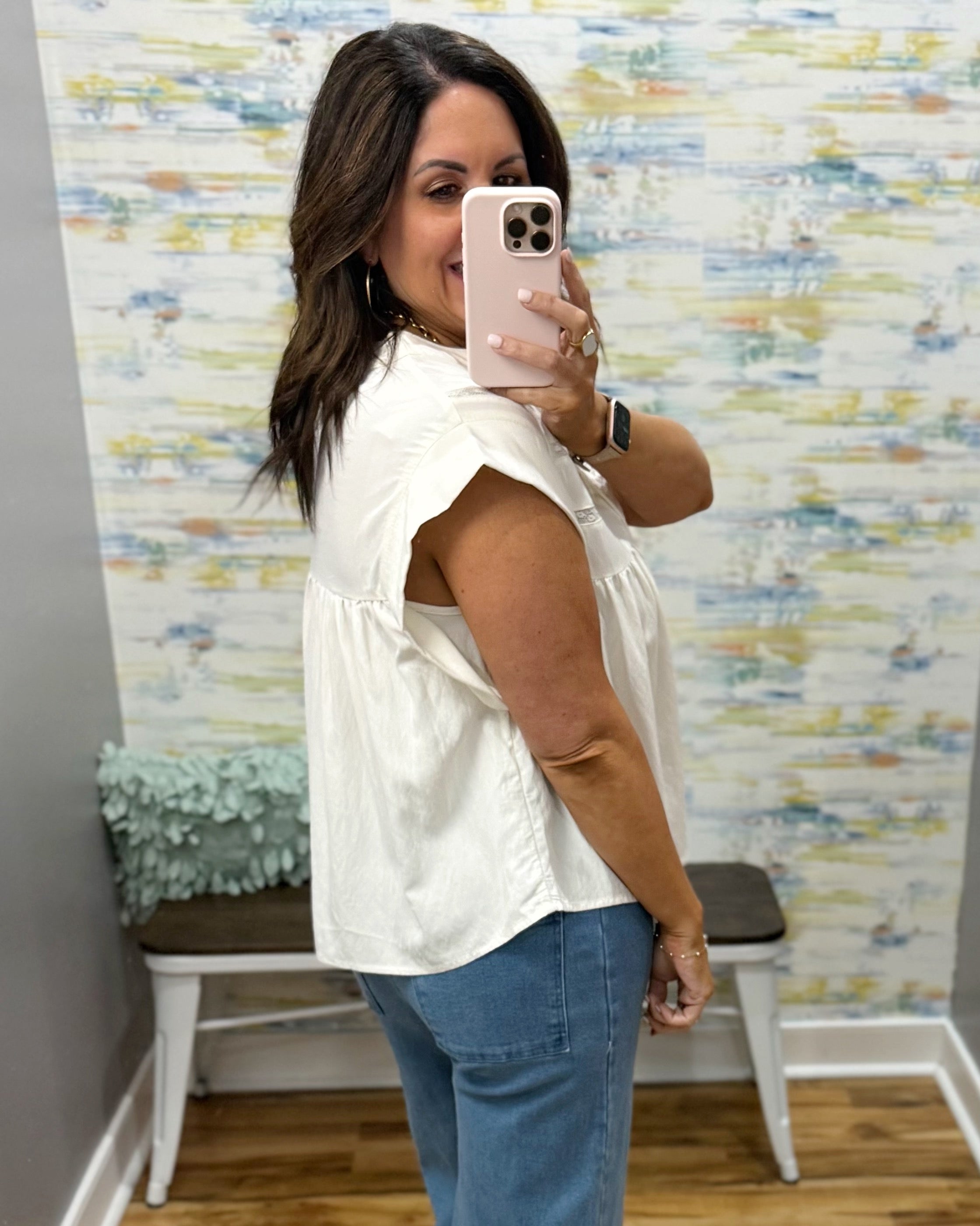 Woman taking a mirror selfie wearing a white sleeveless top and blue jeans in front of a patterned wall.