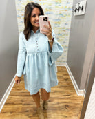 A woman takes a mirror selfie wearing a light blue long-sleeve button-front dress with a gathered waist and flared bell sleeves, paired with tan ankle boots, in an indoor hallway with wood floors.