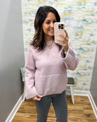 A woman takes a mirror selfie wearing a light pink ribbed knit sweater with front pockets and gray pants in an indoor room with patterned wallpaper.