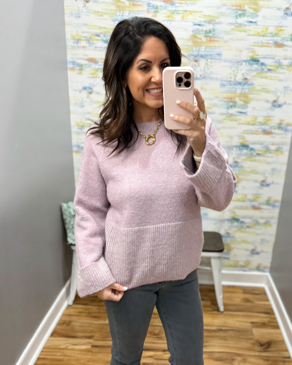 A woman takes a mirror selfie wearing a light pink ribbed knit sweater with front pockets and gray pants in an indoor room with patterned wallpaper.