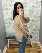 A woman takes a mirror selfie wearing a beige knit sweater with white contrast trim and blue jeans in a room with patterned wallpaper and a bench.