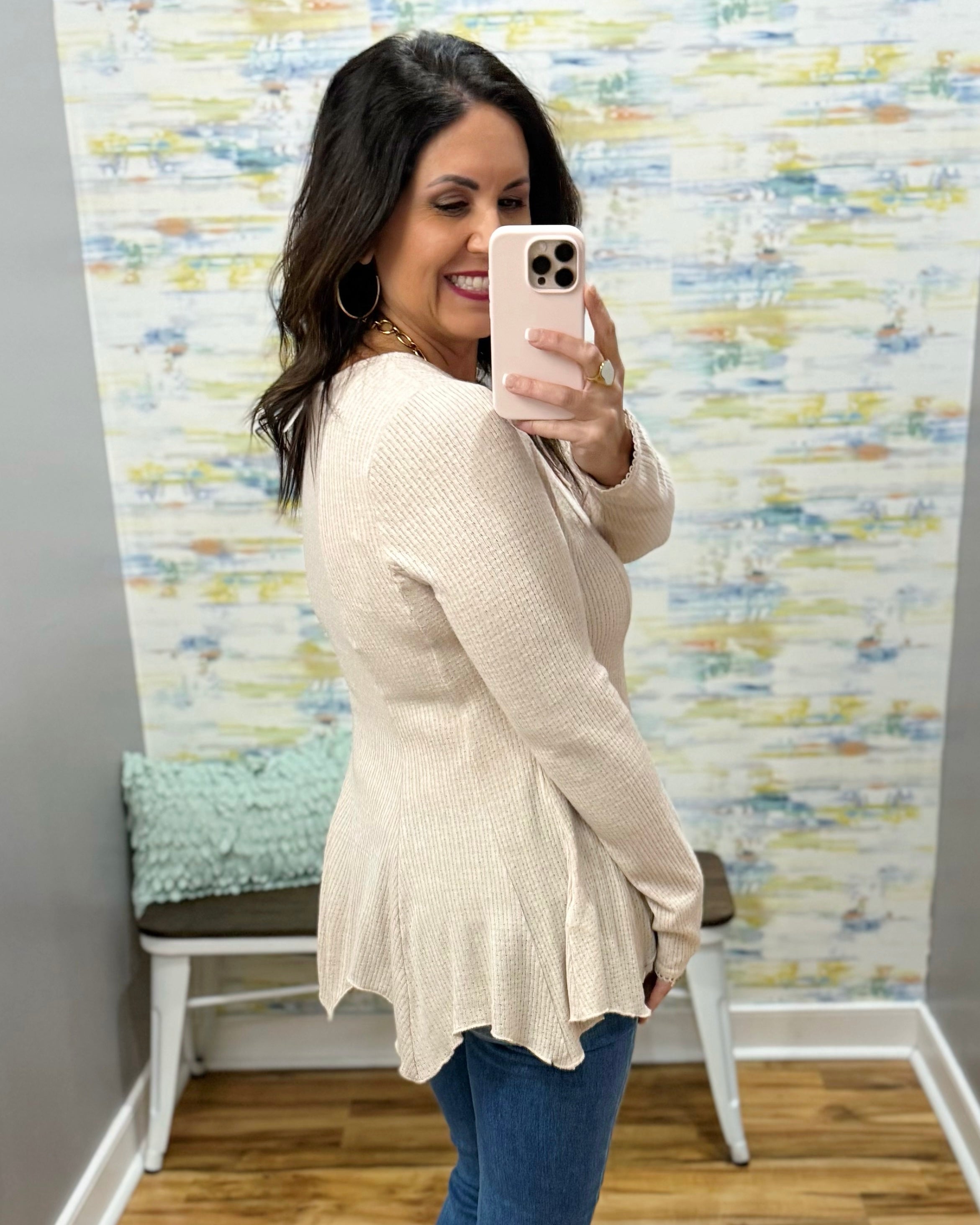 Woman taking a mirror selfie wearing a beige cardigan and blue jeans in front of a multicolored wall.