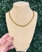 A gold chain necklace displayed on a beige burlap jewelry bust, with a hand holding the base of the bust against a green leafy background.