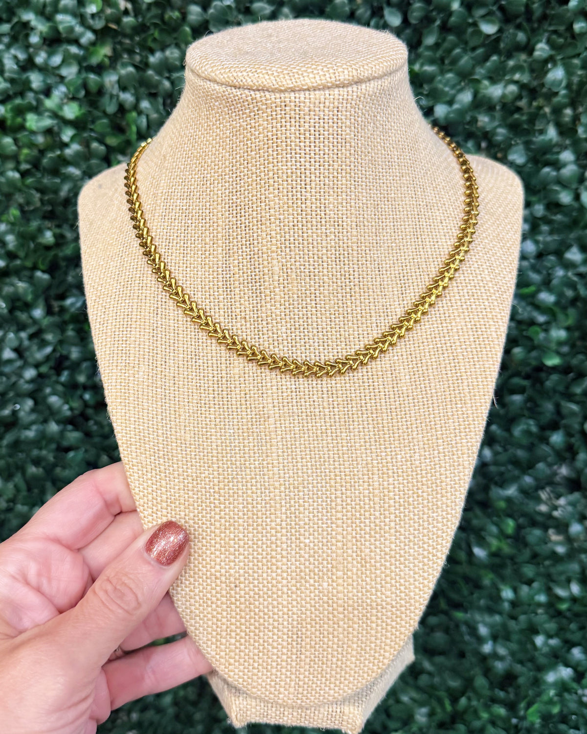 A gold chain necklace displayed on a beige burlap jewelry bust, with a hand holding the base of the bust against a green leafy background.