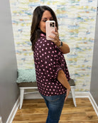 A woman takes a mirror selfie wearing a dark burgundy button-up blouse with pink polka dots and blue jeans, standing in a room with a patterned wall and wooden floor.