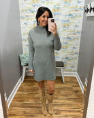 A woman takes a mirror selfie in a fitting room wearing a sage green mock neck knit sweater dress and beige knee-high boots.