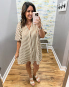 A woman takes a mirror selfie in a fitting room wearing a short-sleeve V-neck mini dress with vertical brown and cream stripes, paired with white lace-up sandals.