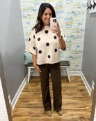 A woman takes a mirror selfie in a fitting room wearing a cream knit top with black polka dots and dark brown wide-leg pants.