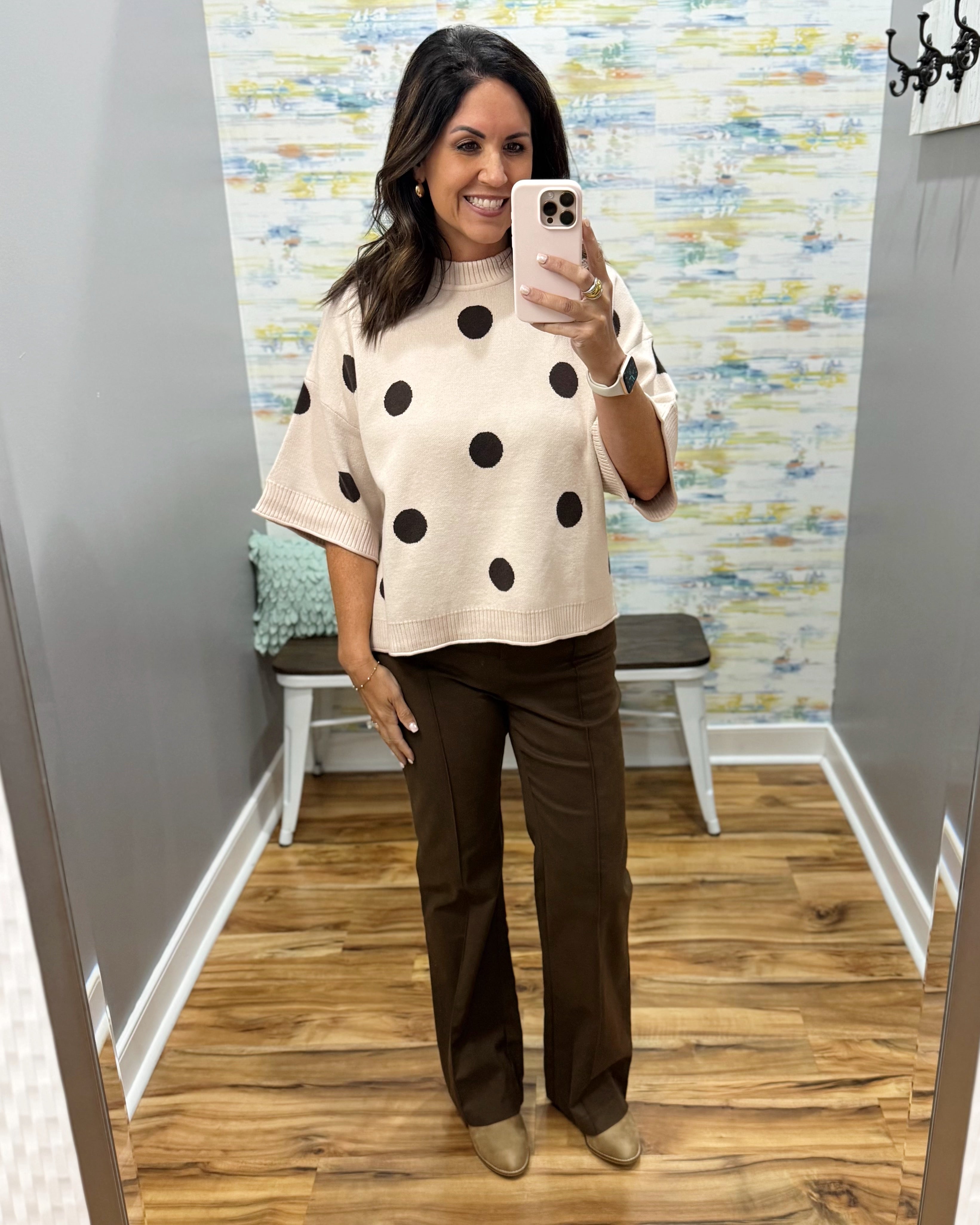 A woman takes a mirror selfie in a fitting room wearing a cream knit top with black polka dots and dark brown wide-leg pants.