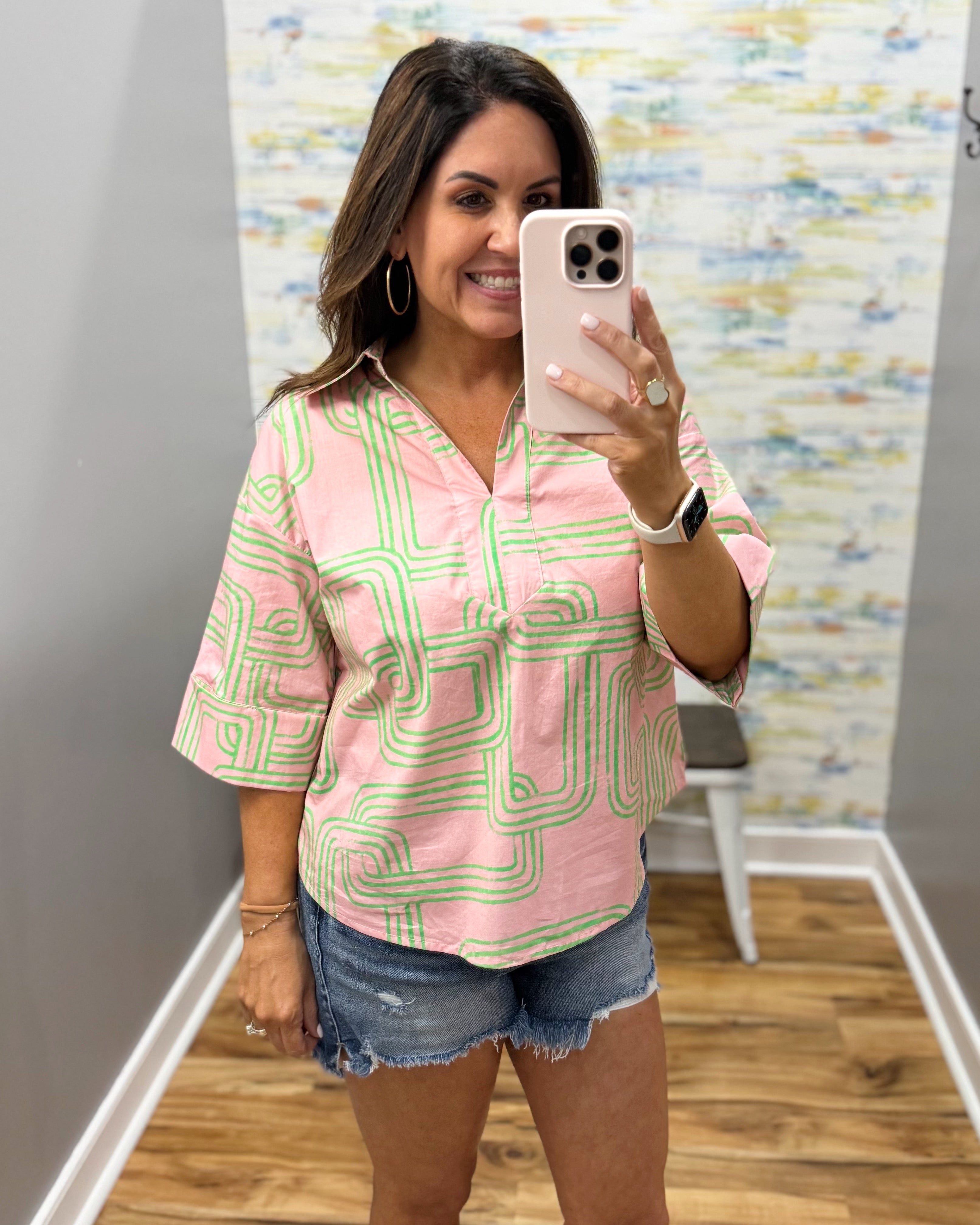 Woman taking a mirror selfie wearing a pink and green patterned shirt and denim shorts.