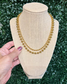 Chansuttpearls double-strand gold beaded necklace displayed on a beige fabric-covered jewelry bust, with a hand holding the left side of the bust against a green leafy background.
