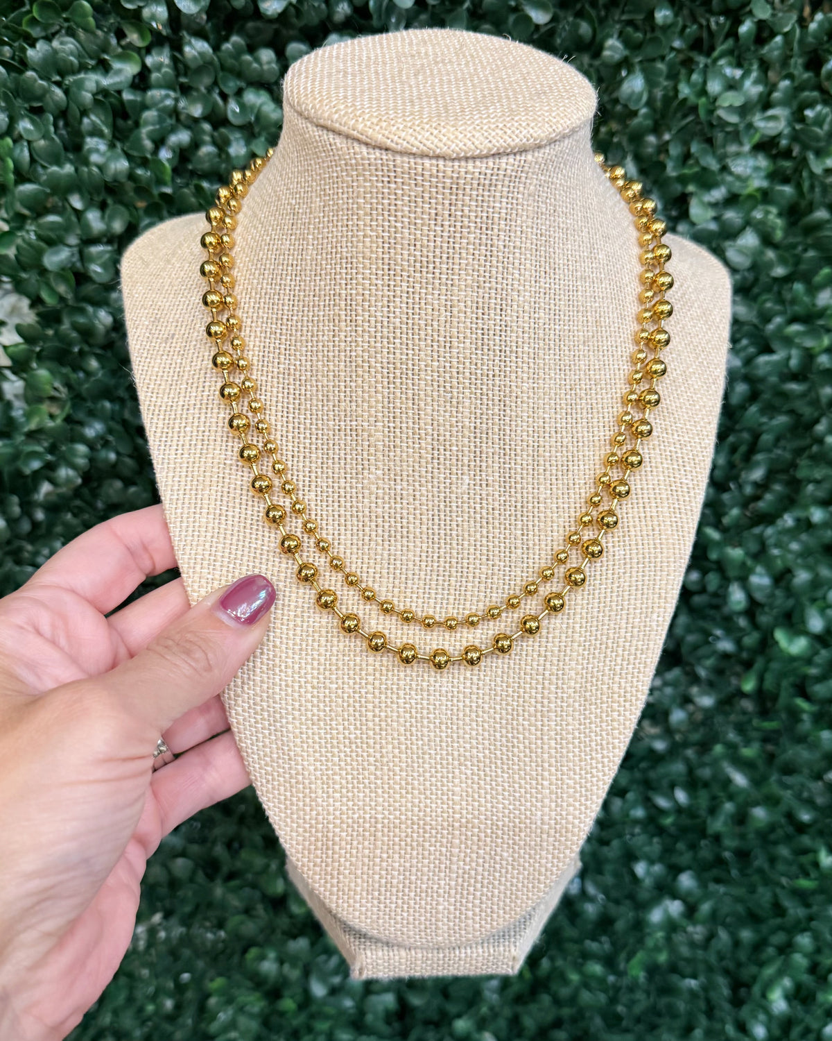Chansuttpearls double-strand gold beaded necklace displayed on a beige fabric-covered jewelry bust, with a hand holding the left side of the bust against a green leafy background.