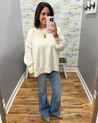 A woman takes a mirror selfie wearing a cream-colored, oversized long-sleeve knit sweater and blue flared jeans in a fitting room with wood floors and gray walls.
