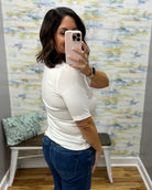 A woman taking a mirror selfie in a fitting room wearing a fitted white short-sleeve top and blue jeans.