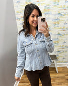 A woman takes a mirror selfie wearing a light-wash blue denim cropped jacket and dark brown pants in an indoor setting with a patterned wall and wood floor.