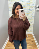 A woman taking a mirror selfie in an indoor hallway, wearing a brown ribbed knit long-sleeve sweater and blue jeans.