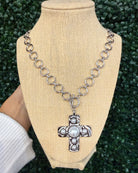 A silver-toned chain necklace with circular links and a cross pendant featuring a central white pearl and small clear stones, displayed on a beige fabric mannequin bust with a hand holding the left side against a green textured background.