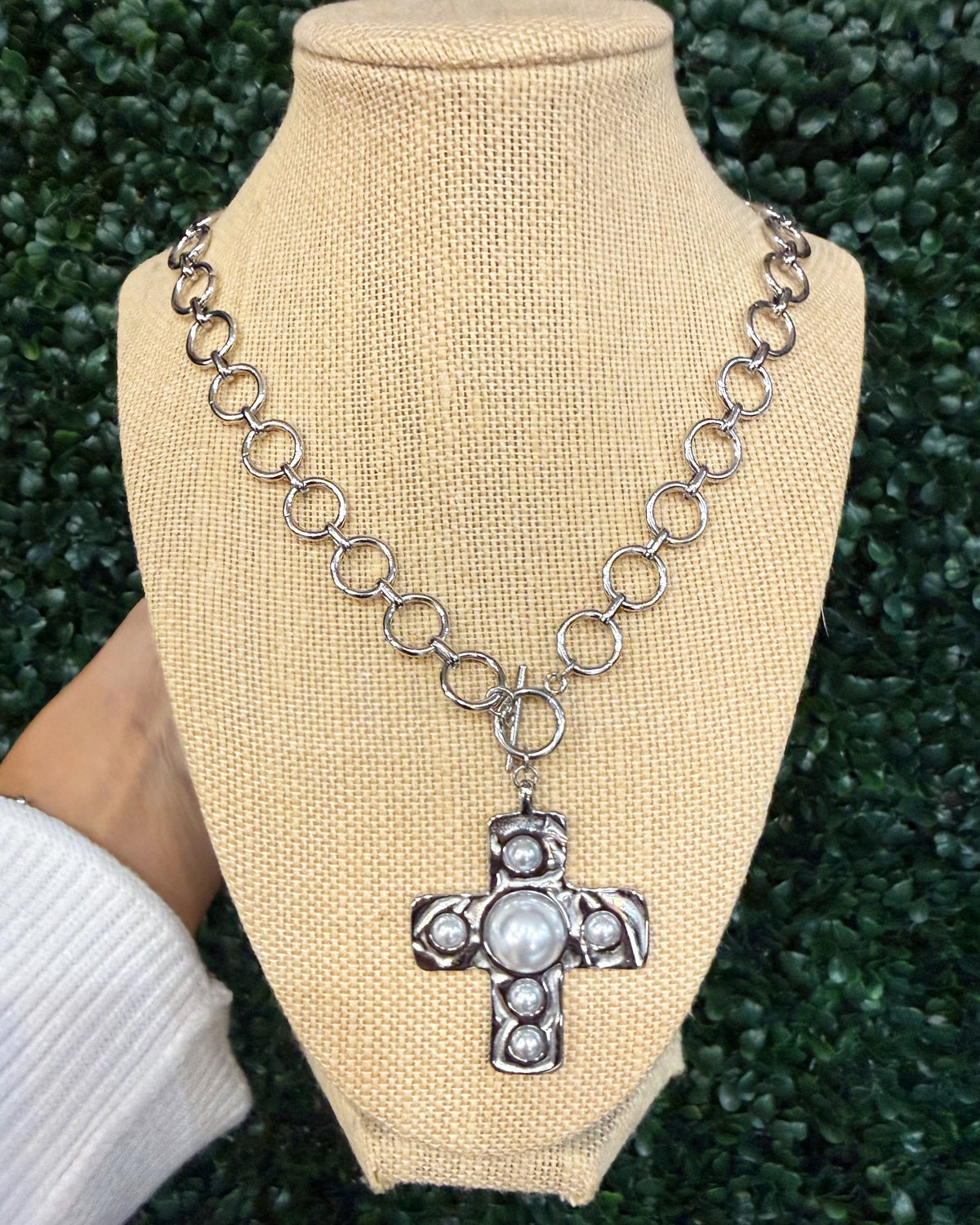 A silver-toned chain necklace with circular links and a cross pendant featuring a central white pearl and small clear stones, displayed on a beige fabric mannequin bust with a hand holding the left side against a green textured background.