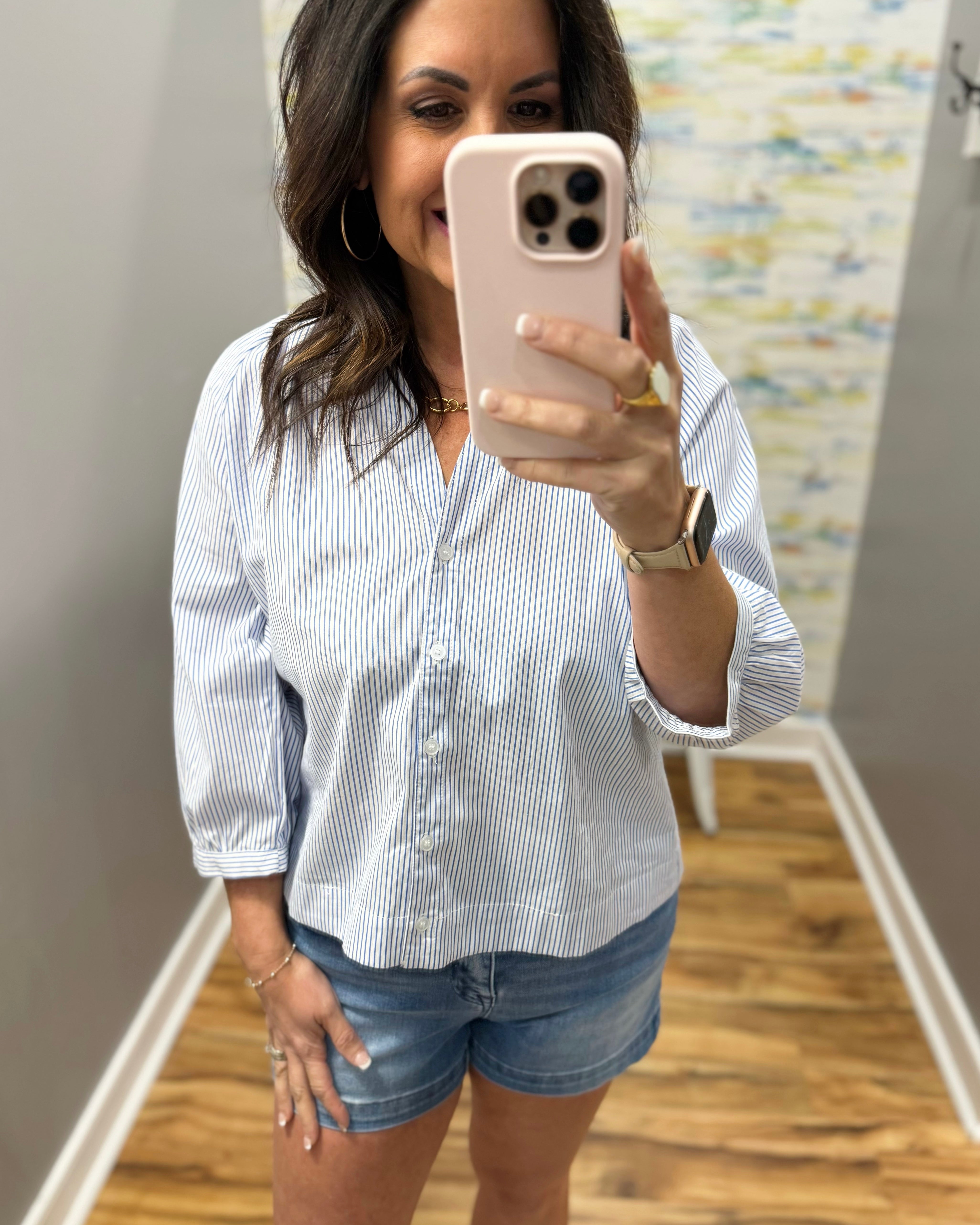 Woman taking a mirror selfie wearing a striped shirt and denim shorts.