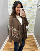 A woman takes a mirror selfie wearing a brown open-front knit cardigan with a white geometric pattern on the lower front, a white top, and blue jeans.