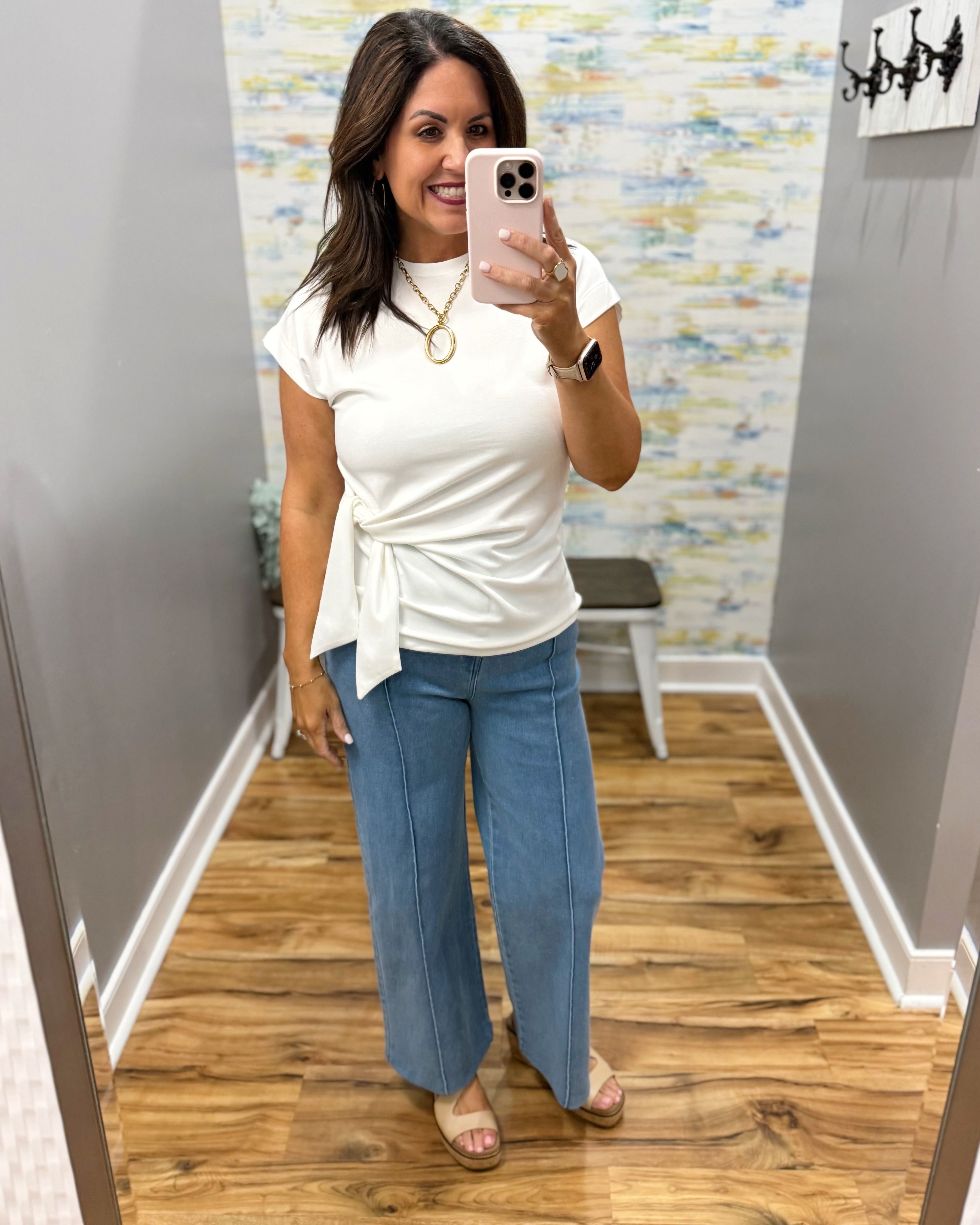 Woman taking a mirror selfie wearing a white top and blue jeans in a room with patterned wallpaper.