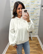 A woman takes a mirror selfie wearing a light heather gray pullover hoodie with a buttoned henley neckline and drawstring, paired with blue jeans in an indoor room with wood floors and patterned wallpaper.
