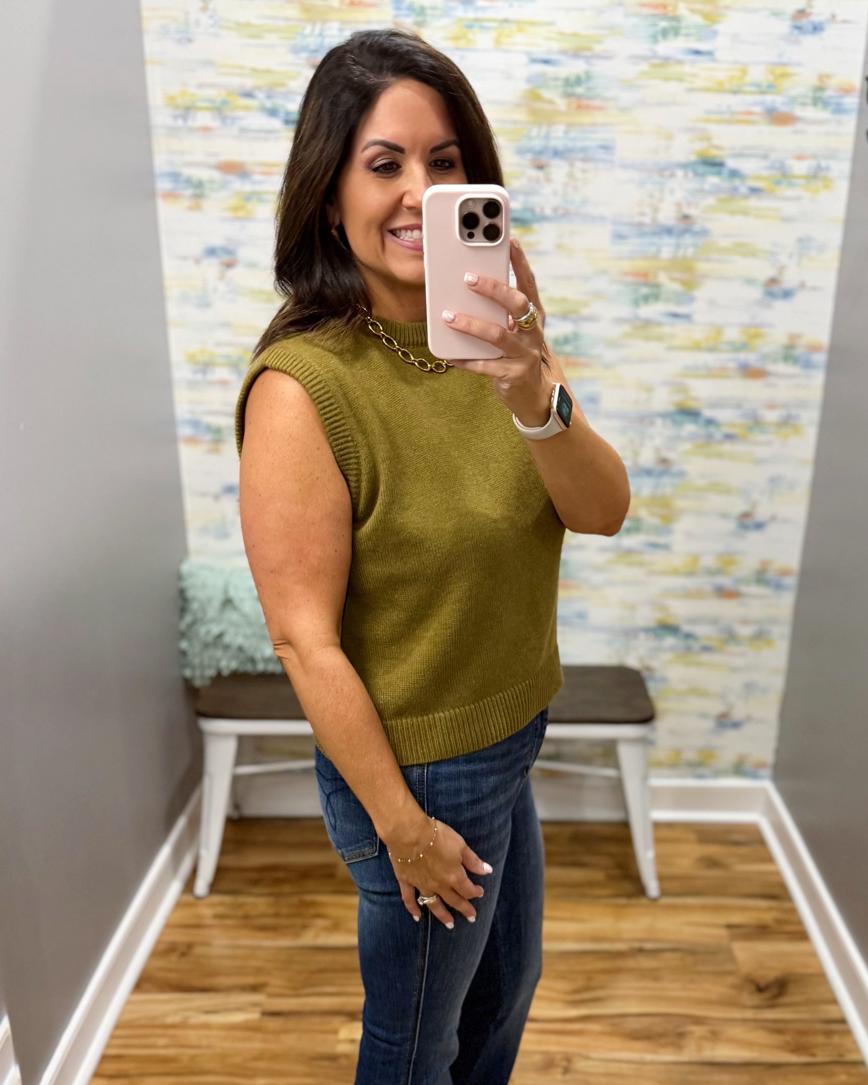 A woman takes a mirror selfie wearing a sleeveless olive green knit sweater vest and blue jeans in a fitting room with a patterned wallpaper background.