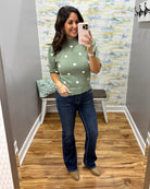 A woman takes a mirror selfie in a fitting room wearing a sage green knit sweater with white polka dots and dark blue flare jeans.