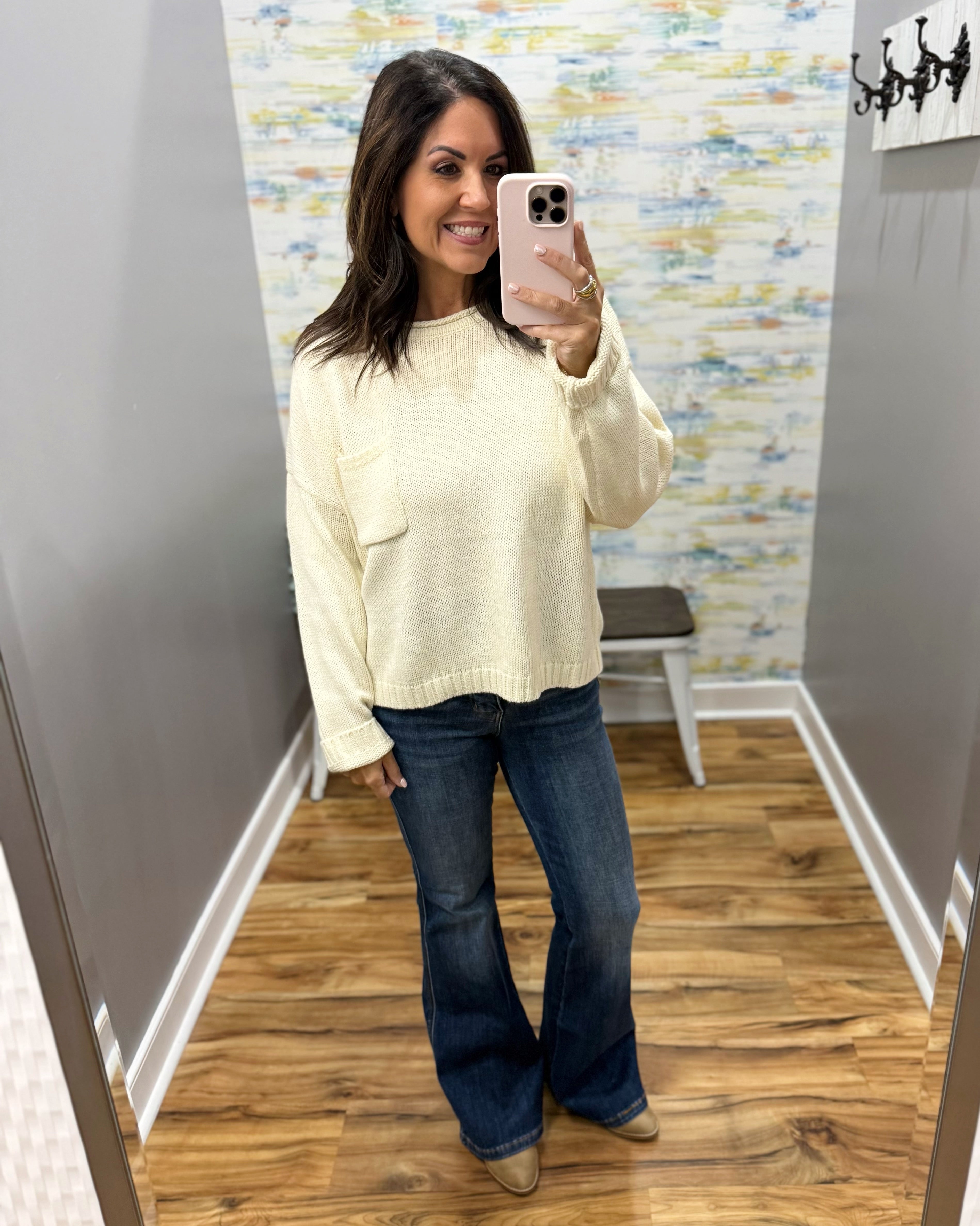 A woman takes a mirror selfie in a fitting room wearing a cream-colored knit sweater and dark blue flare jeans.