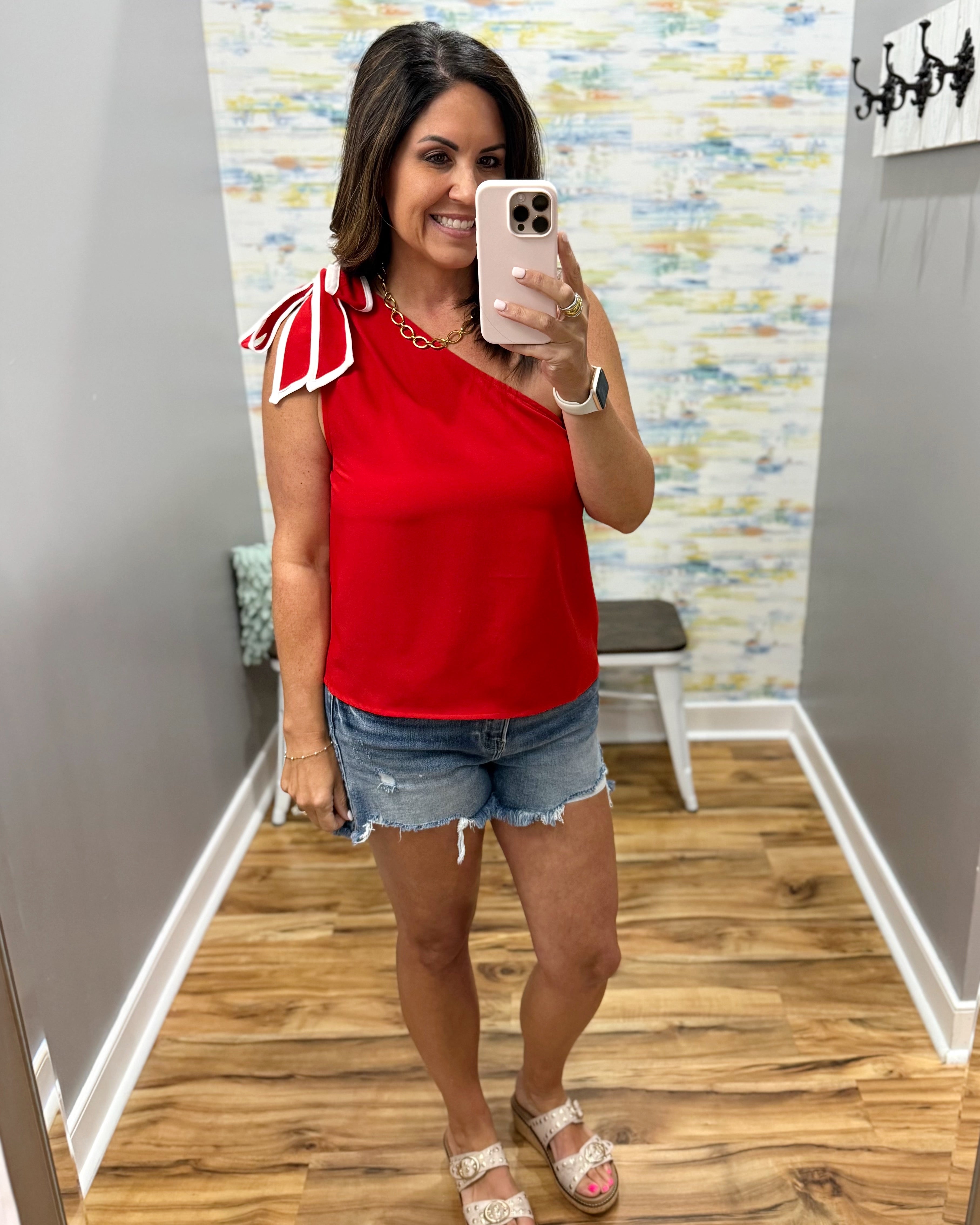 Woman taking a mirror selfie wearing a red top and denim shorts in a room with a colorful wall.