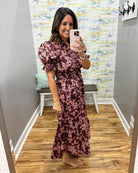 A woman takes a mirror selfie in a fitting room wearing a dark burgundy maxi dress with a pink and white floral pattern, puff sleeves, and a V-neckline.