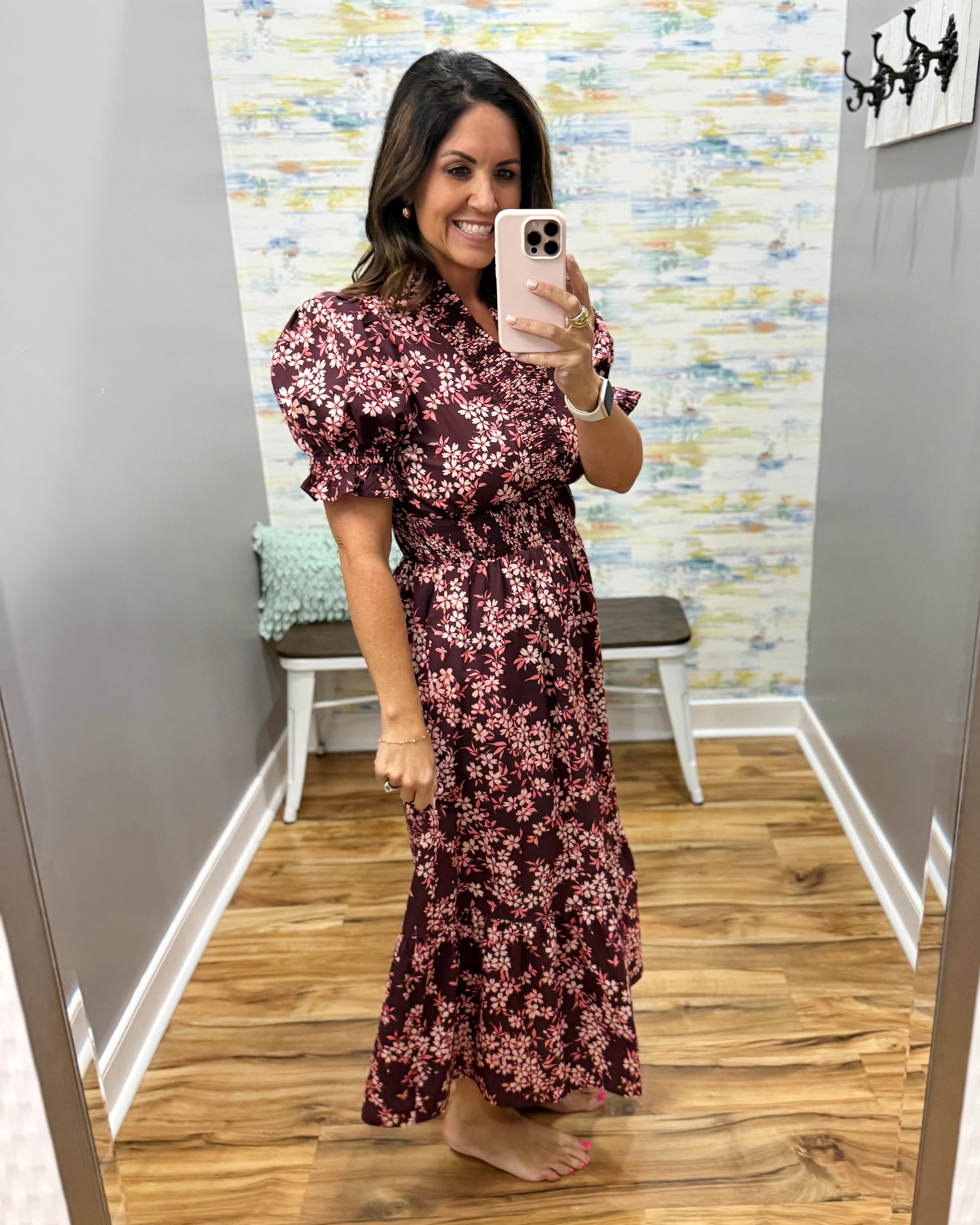 A woman takes a mirror selfie in a fitting room wearing a dark burgundy maxi dress with a pink and white floral pattern, puff sleeves, and a V-neckline.