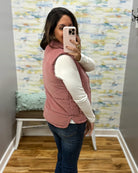 A woman takes a mirror selfie wearing a dusty rose quilted vest over a white long-sleeve shirt and dark blue jeans, standing in a room with patterned wallpaper and a wooden floor.