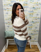 A woman takes a mirror selfie wearing a cream and brown horizontally striped knit sweater, blue jeans, and a light pink phone case in a room with patterned wallpaper.