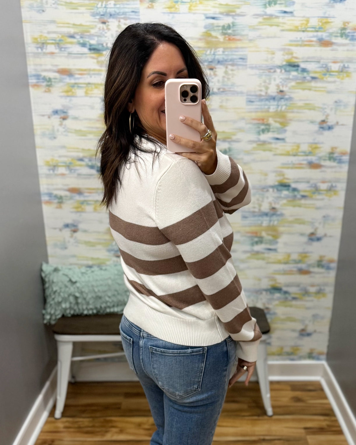 A woman takes a mirror selfie wearing a cream and brown horizontally striped knit sweater, blue jeans, and a light pink phone case in a room with patterned wallpaper.