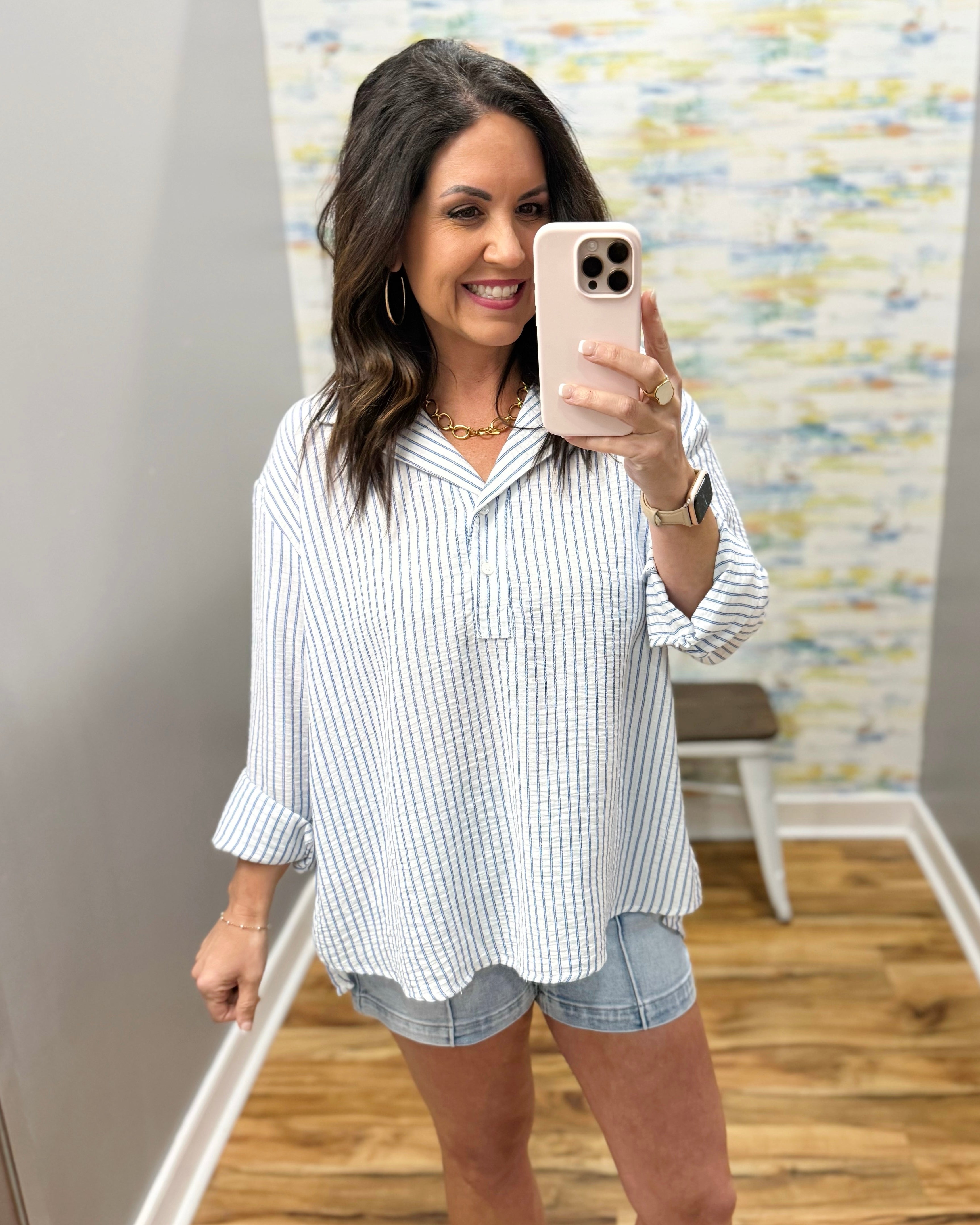 Woman taking a mirror selfie wearing a striped shirt and denim shorts in a room with a patterned wall.