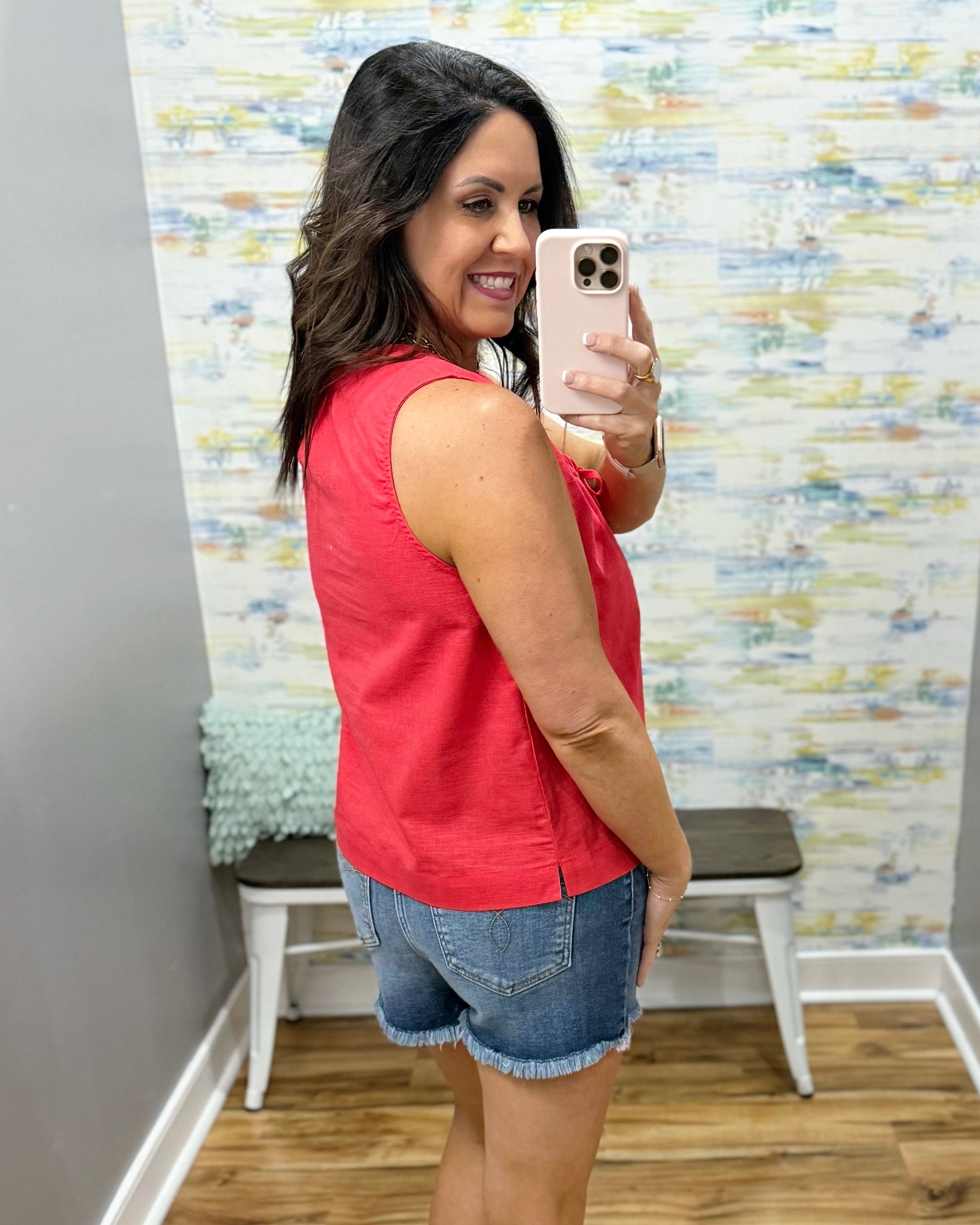 Woman taking a mirror selfie wearing a red sleeveless top and denim shorts in a room with a textured wall.