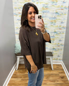 A woman takes a mirror selfie wearing a loose-fitting, dark brown short-sleeve top and light blue jeans in a room with wood floors and a patterned wallpaper background.