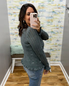 A woman takes a mirror selfie in a fitting room wearing a dark gray ribbed knit sweater with small white embroidered details, paired with blue jeans.